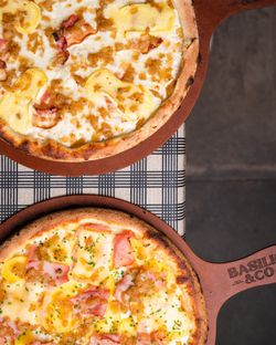 Deux pizzas artisanales sur des planches en bois avec la marque Basilic & Co, posées sur une table à carreaux.