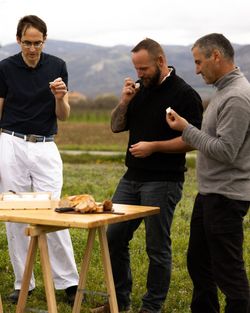 Trois hommes debout dans un champ dégustent des morceaux de fromage autour d'une table en bois avec du pain.