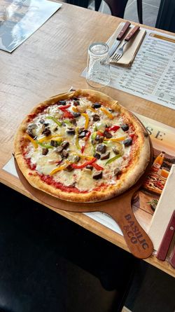 Pizza artisanale aux légumes variés sur une table en bois avec couverts, menu et verre retourné au restaurant Basilic & Co