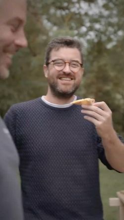 Homme souriant tenant une part de pizza en extérieur, ambiance conviviale avec une autre personne floue en premier plan.