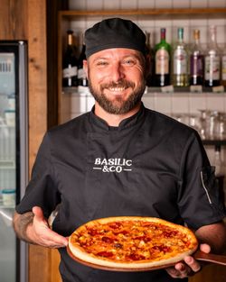 Pizzaiolo en uniforme noir tenant une pizza artisanale cuite au four dans un restaurant avec bouteilles en arrière-plan.