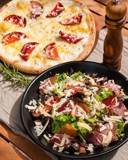 Pizza artisanale garnie de tranches de charcuterie et salade fraîche avec fromage râpé dans un bol noir.
