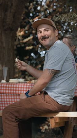 Homme assis à une table en bois avec nappe à carreaux, mangeant à l'extérieur sous un arbre, ambiance décontractée.