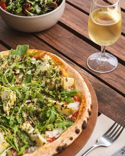 Pizza artisanale avec roquette, artichauts et pesto, accompagnée d'un verre de vin blanc et d'une salade fraîche.