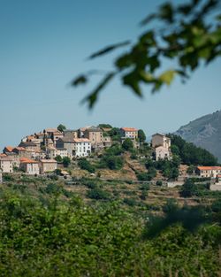 Vue sur un village perché avec des maisons anciennes entourées de végétation et une colline en arrière-plan.