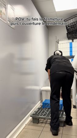 Personne en tenue noire dans un espace de travail étroit avec palettes et caisses, ambiance cuisine.