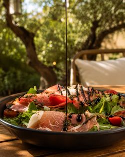 Salade fraîche avec jambon cru, tomates, roquette, et filet de sauce balsamique, servie en extérieur au restaurant.