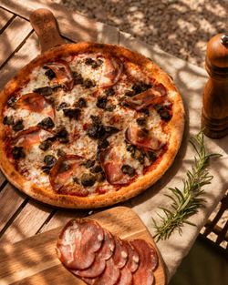 Pizza artisanale garnie de fromage, charcuterie, et champignons, présentée sur une table en bois avec herbes fraîches.