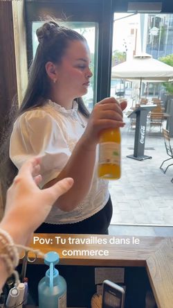Serveuse tenant une bouteille de boisson dans un restaurant avec terrasse extérieure visible par la fenêtre.