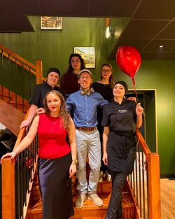 Équipe de six personnes posant sur un escalier en bois dans un restaurant avec mur vert. Une personne tient un ballon rouge.