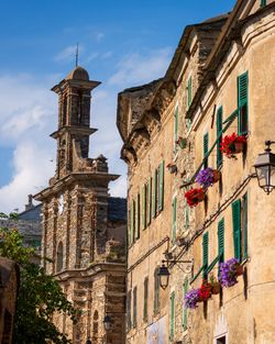 Façades anciennes avec volets verts et fleurs, et clocher en pierre sous un ciel bleu avec quelques nuages.