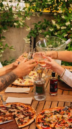Plusieurs pizzas artisanales posées sur une table en bois avec des mains portant un toast au vin blanc.