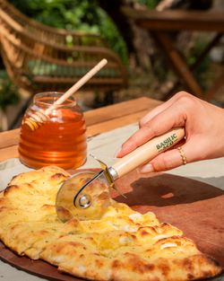 Découpe d'une pizza artisanale avec roulette à pizza sur planche en bois, pot de miel à côté sur table en extérieur.