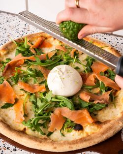 Pizza artisanale garnie de roquette, saumon fumé et une boule de burrata au centre, zeste de citron râpé dessus.
