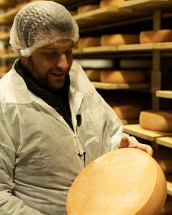 Employé en tenue de protection tenant une grande meule de fromage dans une fromagerie avec étagères remplies de fromages.