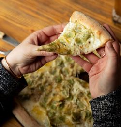 Pizza artisanale à base de pâte épaisse avec garniture crémeuse et légumes verts, tenue par deux mains.
