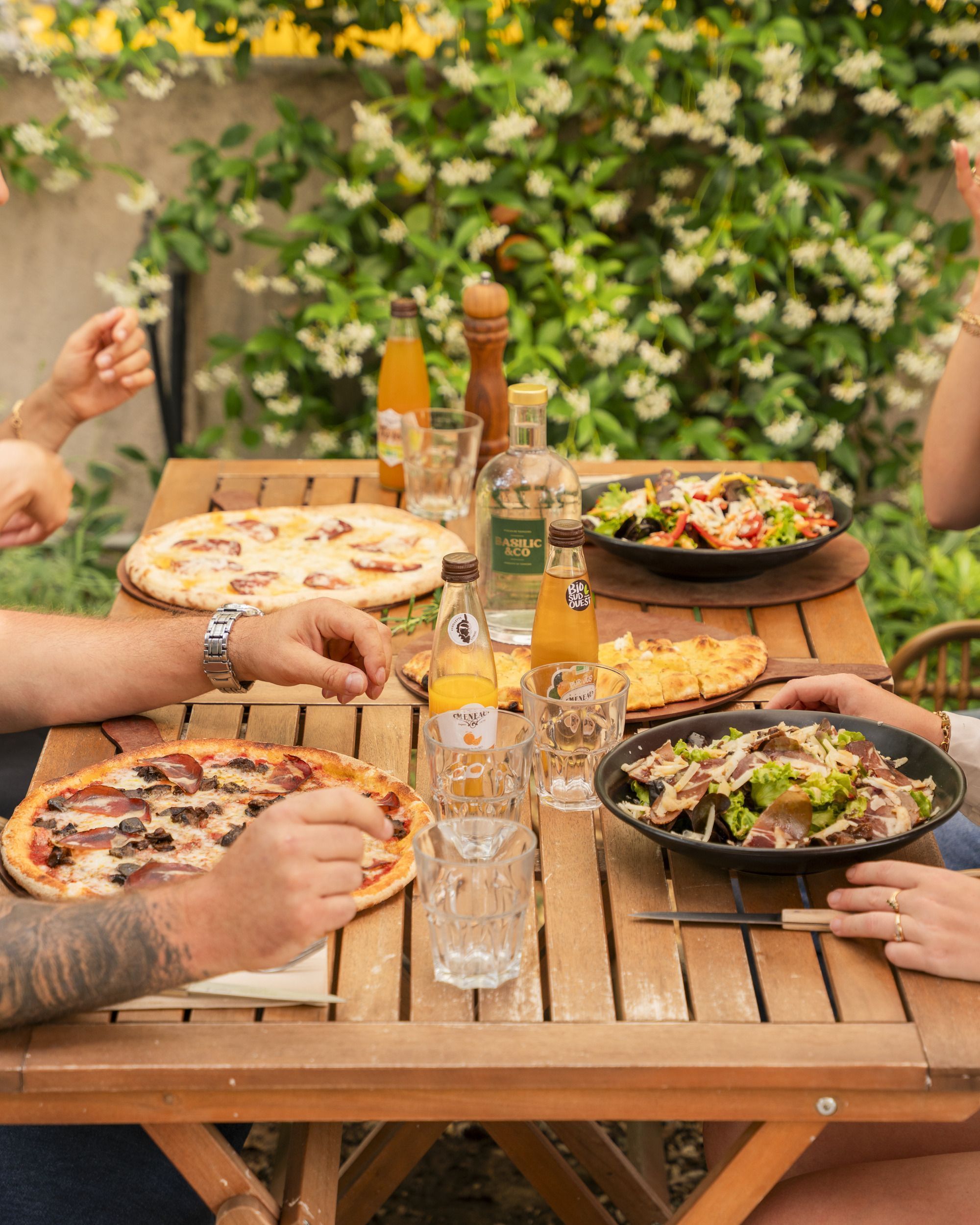 Pizza artisanale et salades servies sur une table en bois en extérieur avec des boissons et des verres. Repas convivial.