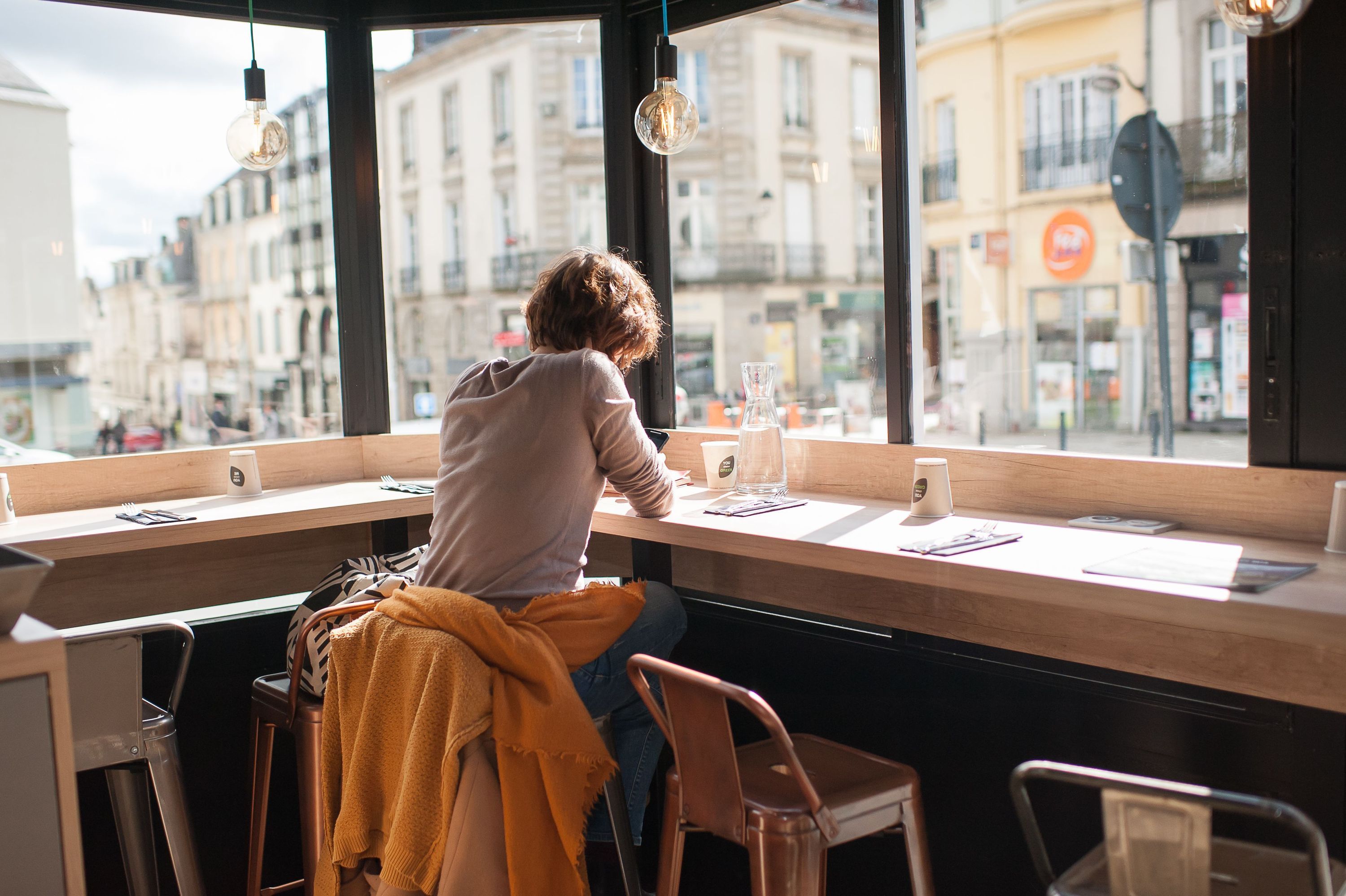 Espace de restauration avec bar en bois et grandes fenêtres donnant sur la rue, éclairage par ampoules suspendues.