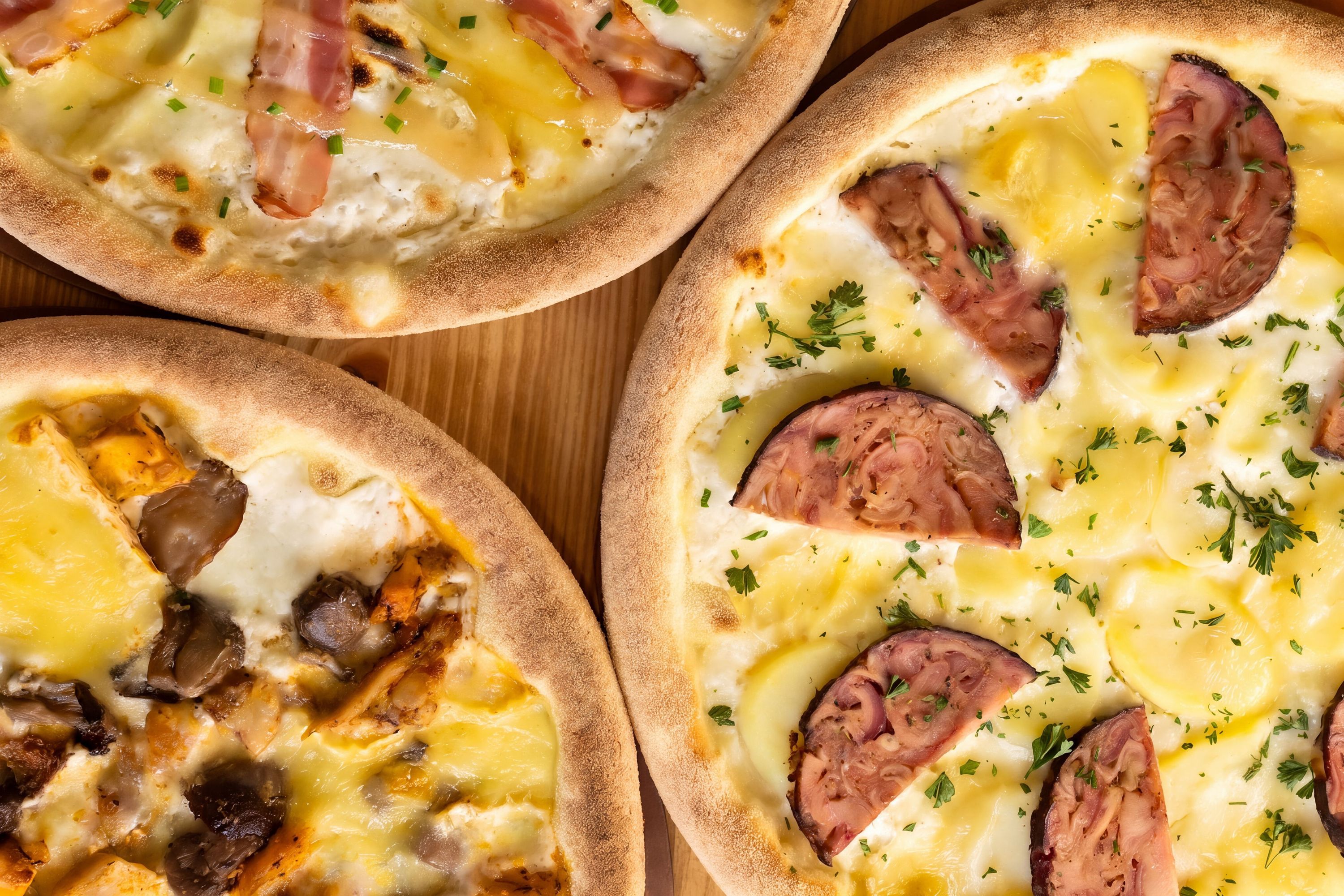 Trois pizzas artisanales avec différentes garnitures sur un plateau en bois, proposées à Basilic & Co.