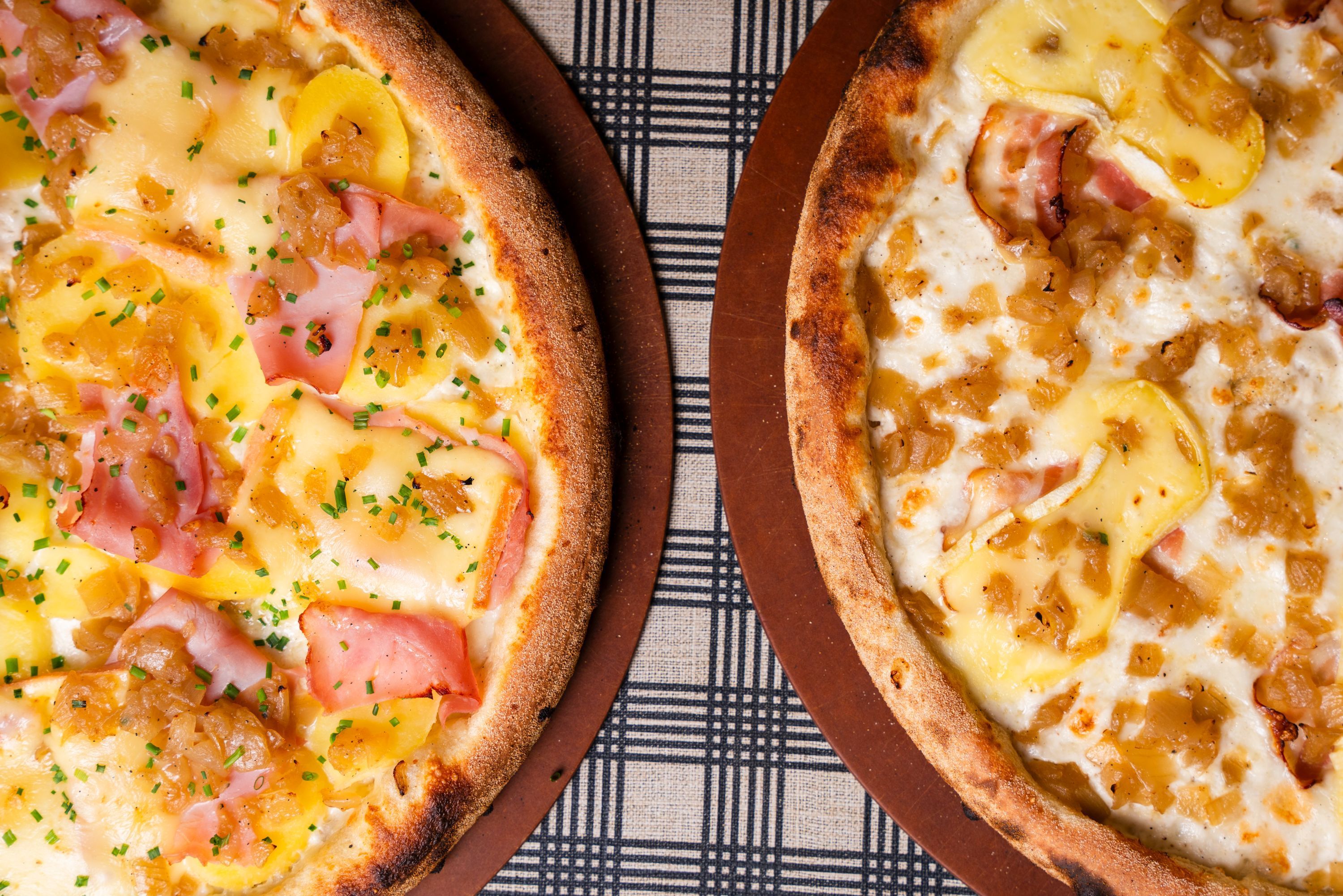 Deux pizzas artisanales garnies de fromage, jambon et oignons caramélisés, présentées sur une table à carreaux.