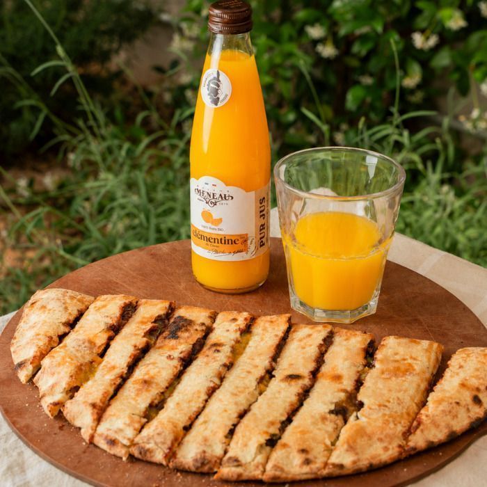 Pizza artisanale coupée en tranches et bouteille avec verre de jus de clémentine sur table en bois.