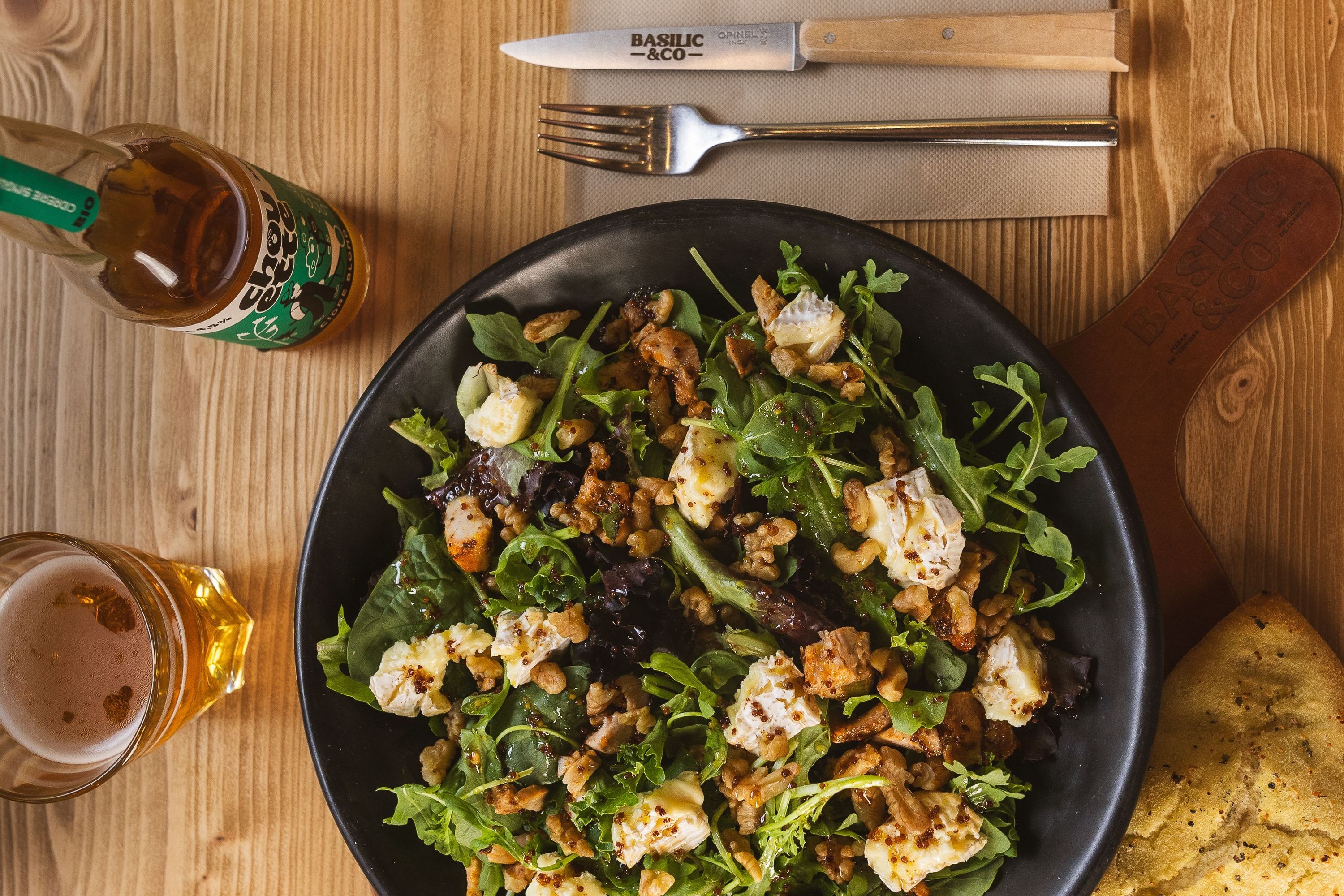 Salade composée avec fromage, noix et roquette servie sur une assiette noire avec une bouteille de bière et un verre.