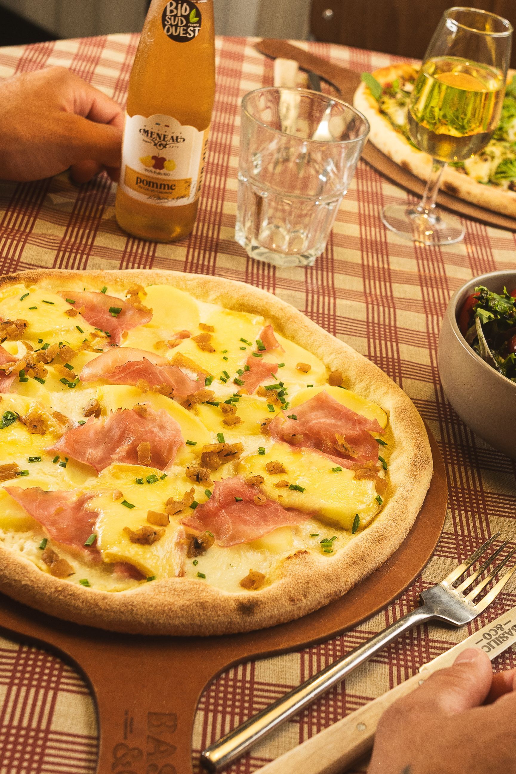 Pizza artisanale avec jambon et fromage sur table à carreaux avec boisson et salade en accompagnement.
