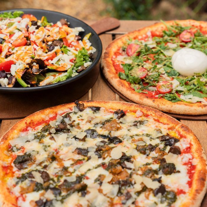 Deux pizzas artisanales avec garnitures variées accompagnées d'une salade fraîche en bol noir sur table en bois.