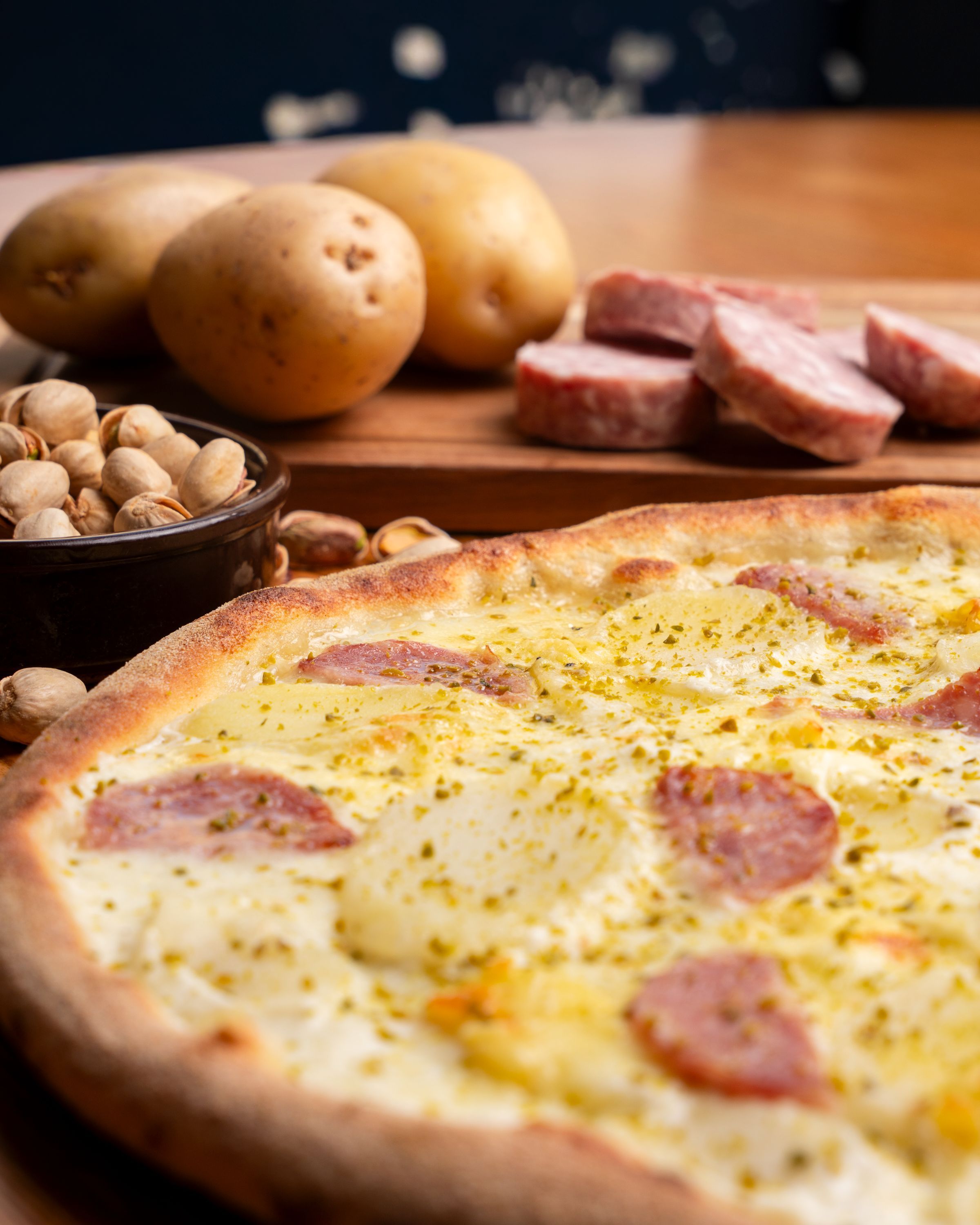 Pizza artisanale avec tranches de saucisse, pommes de terre, et noix sur fond en bois pâle.