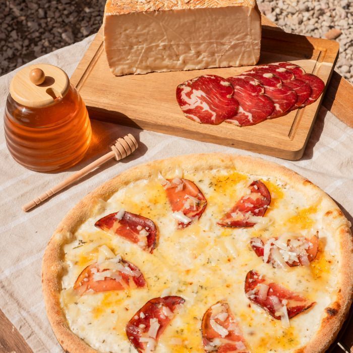 Pizza artisanale garnie de tranches de viande et fromage, accompagnée de miel et fromage sur planche en bois