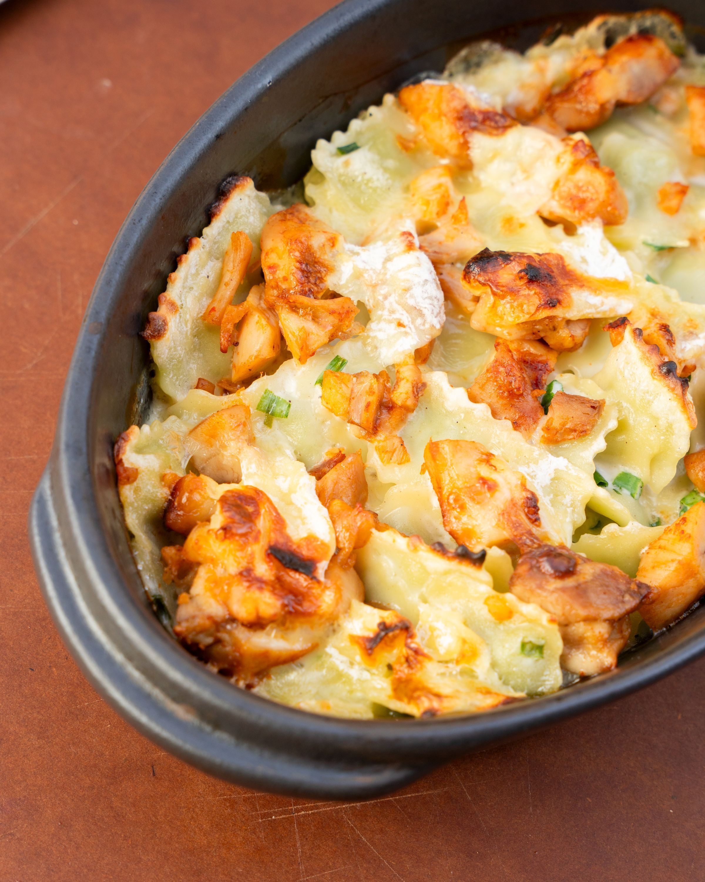 Gratin de ravioles aux morceaux de poulet doré et fromage fondu dans un plat en céramique noire.