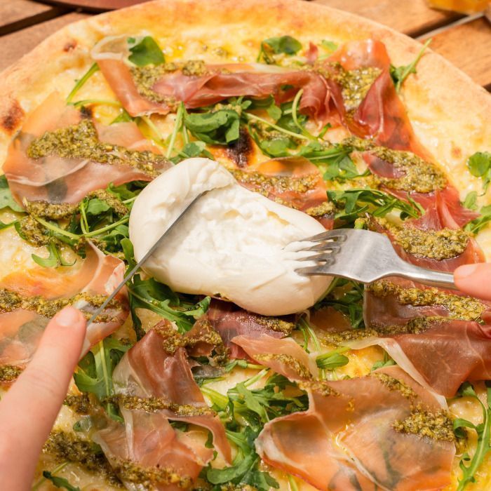 Pizza artisanale garnie de jambon cru, roquette, pesto et mozzarella crémeuse coupée à la main.