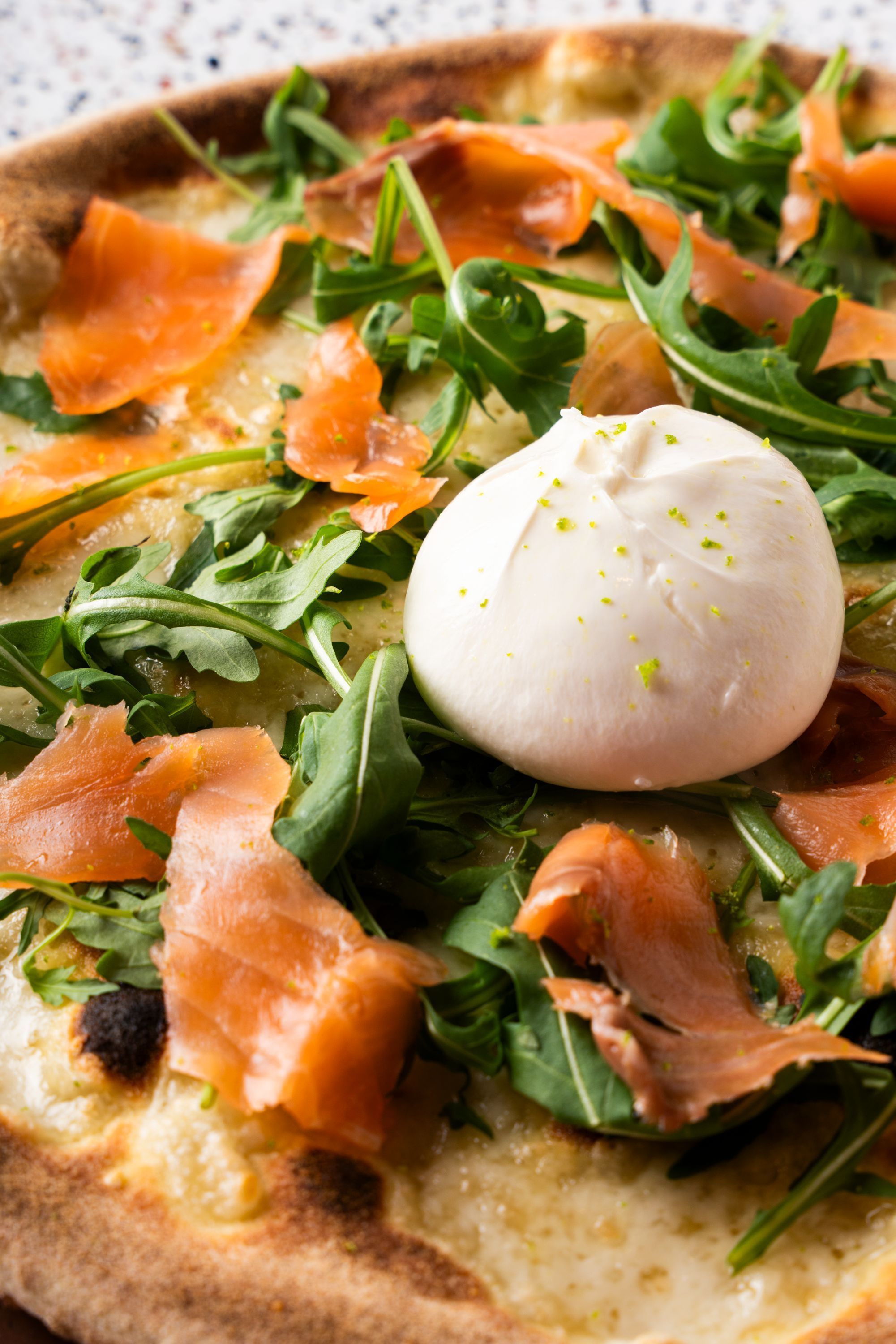 Pizza artisanale garnie de morceaux de saumon fumé, roquette fraîche et une boule de burrata au centre.